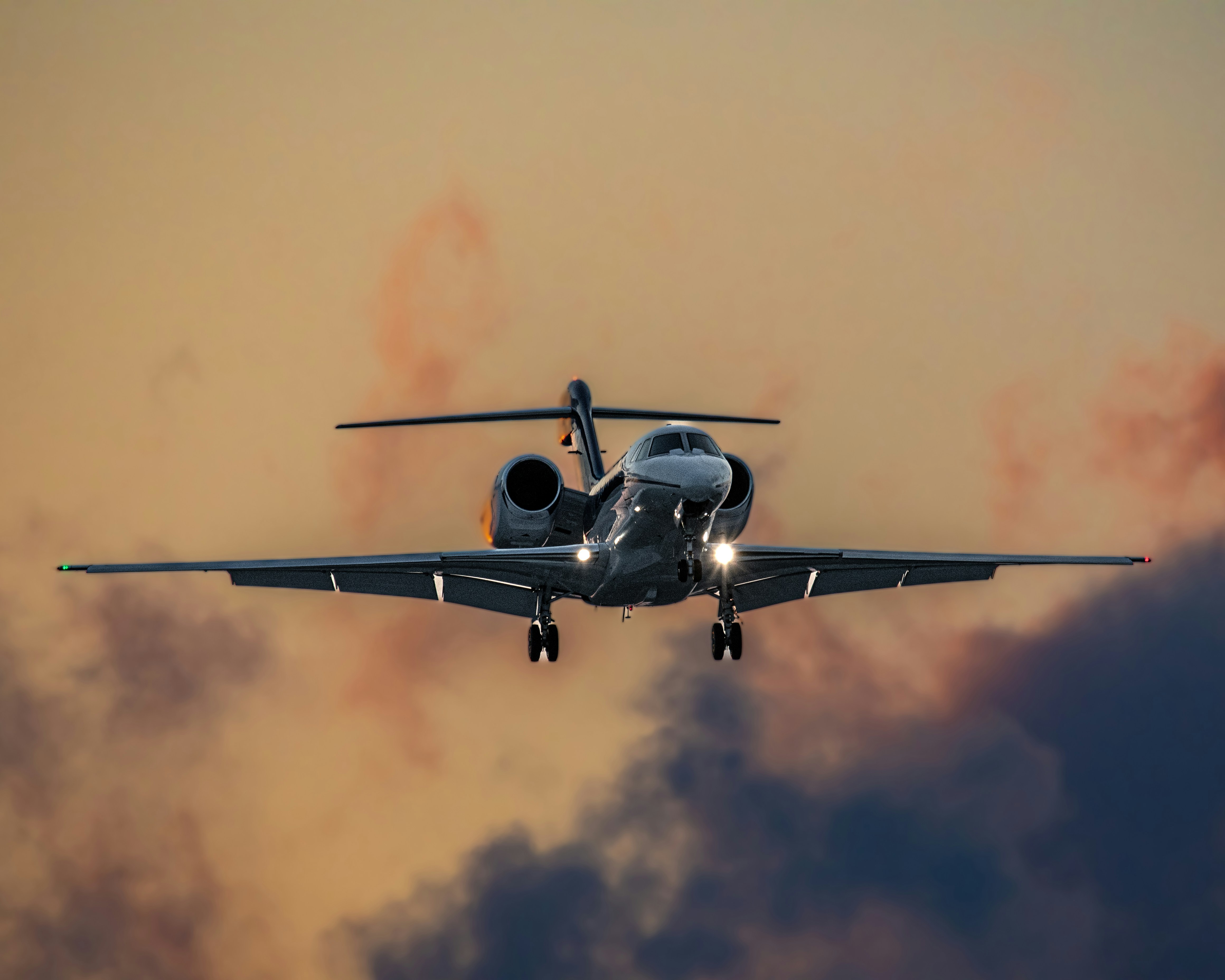 Private jet landing with lights on against a dramatic orange sunset sky.
